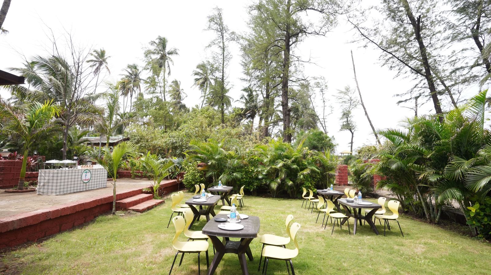 Outdoor dining area with tables and chairs amidst lush greenery and coconut trees - Tranquil Beach Resort, Harihareshwar