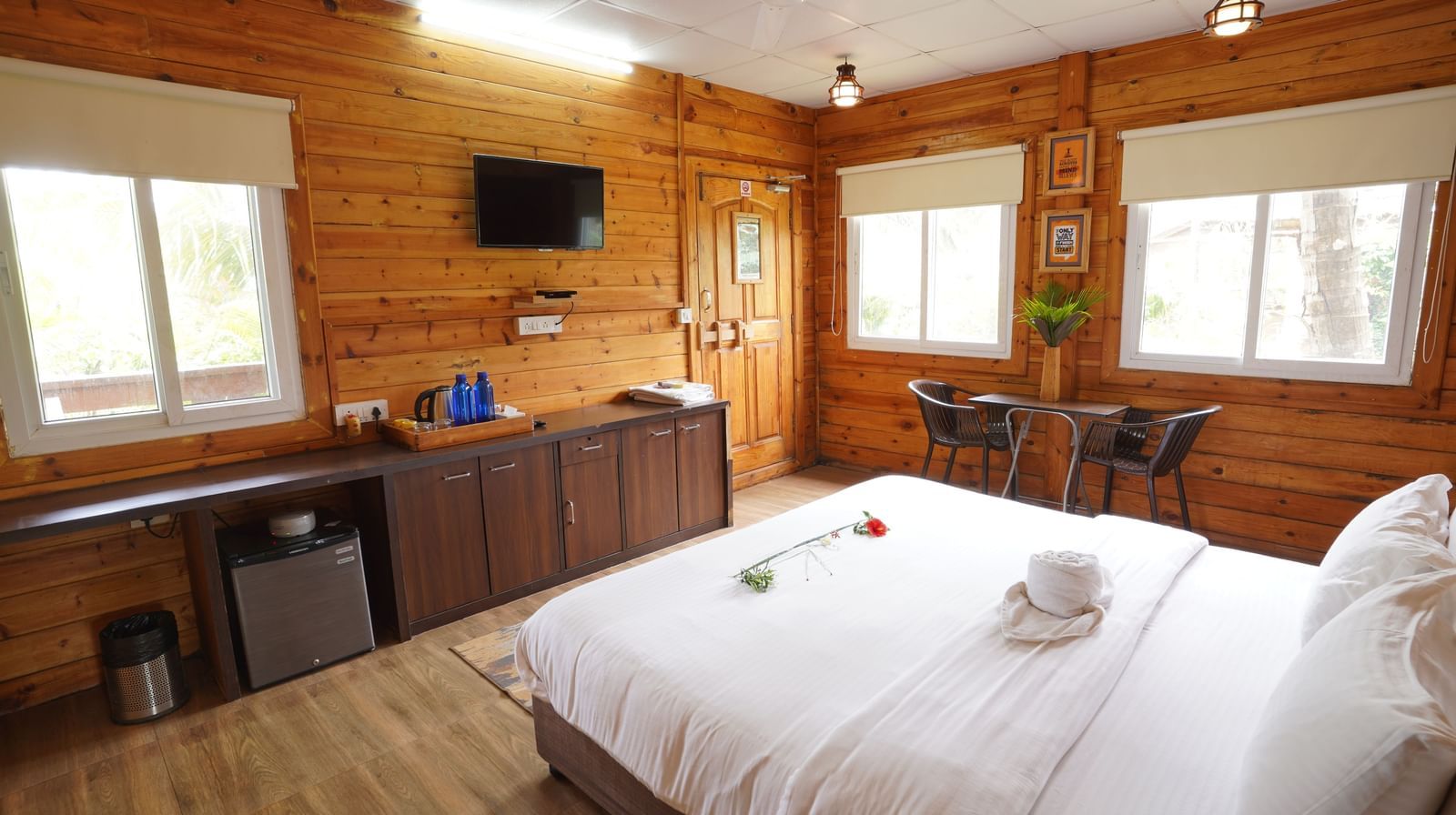 A cosy wooden room with a bed, windows, a mini fridge, a tea and coffee maker, a television, and a small seating area - Tranquil Beach Resort, Harihareshwar