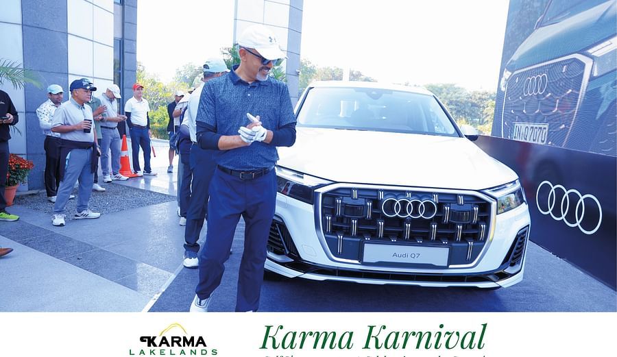 A person holding a microphone standing next to a luxury car, with the Karma Karnival backdrop visible.