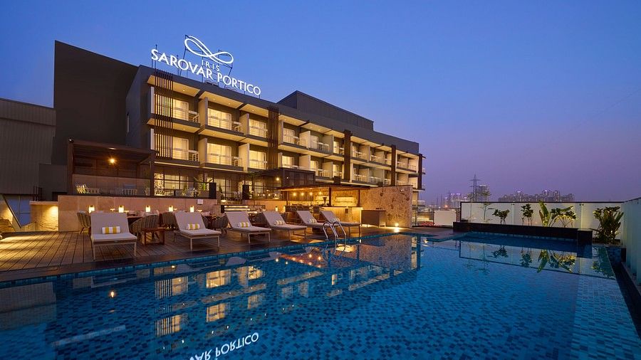 A modern hotel building is illuminated at dusk, featuring a rooftop swimming pool reflecting the hotel's signage and nearby lounge chairs. - Iris Sarovar Portico, Sector 85, Gurugram