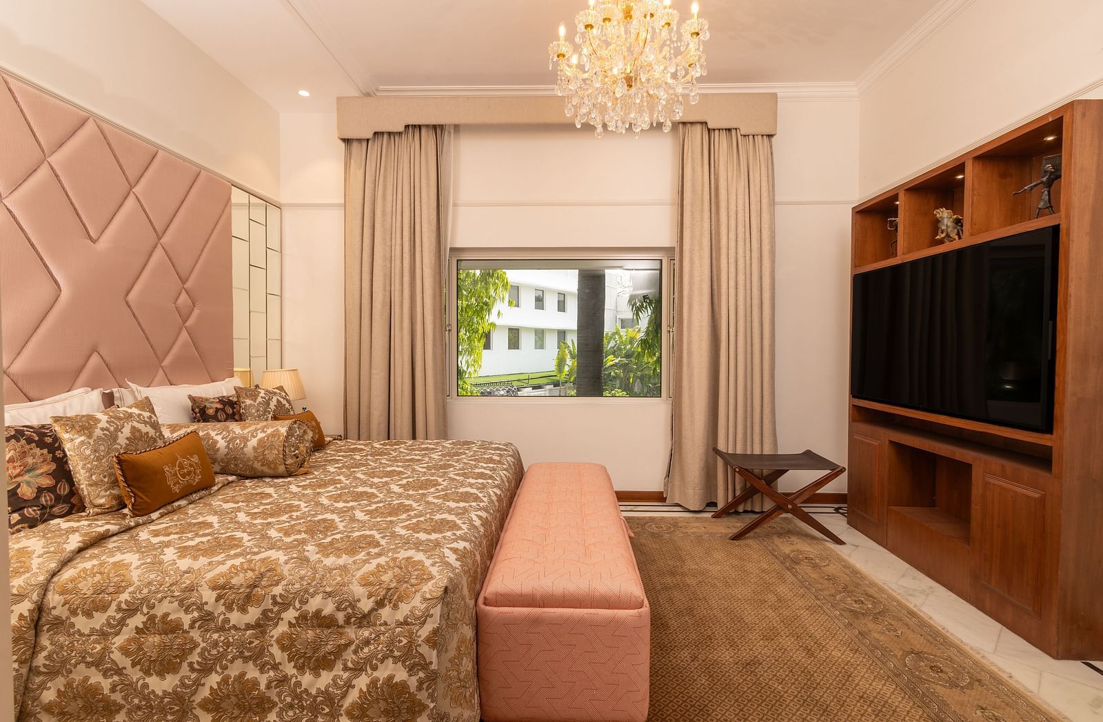 Luxury Suite at The Claridges New Delhi, featuring a cosy bed, wall-mounted TV, chandelier overhead, and a serene view through the window