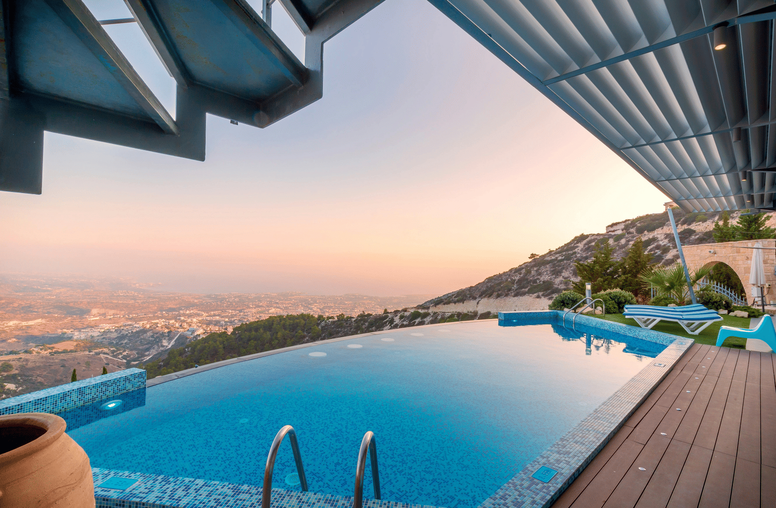 Infinity pool on a hillside with scenic views of the city and mountains at sunset, beside a wooden deck.