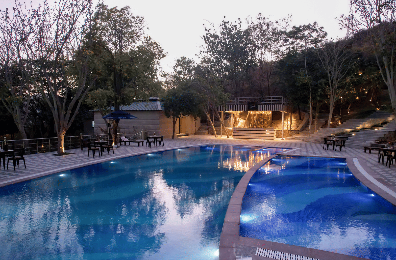 Swimming pool at Essentia Luxury Resort & Spa, Udaipur with tables and lounge areas on the deck surrounded by trees
