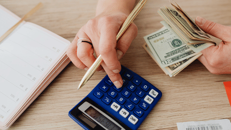 A person uses a calculator beside a notebook while counting US dollar bills on a wooden desk