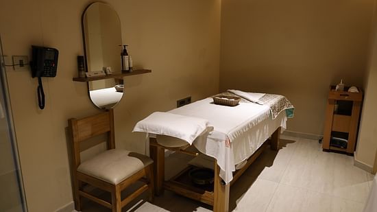 Massage room with a wooden bed covered in white linens and a folded patterned blanket, a chair beside a wall-mounted mirror and shelf with bottles, a wall telephone, and a wooden trolley in the corner under soft ambient lighting - Golden Tulip, Lucknow