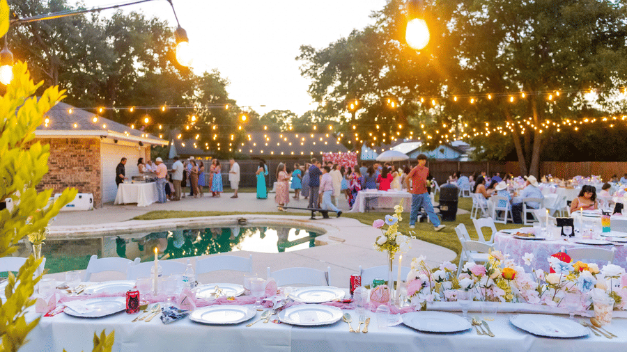 An outdoor gathering taking place near a swimming pool under strings of hanging lights, with a set table in the foreground.