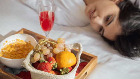 A woman in white robes lying beside a tray of fruits, a yellow dish, and a glass filled with red wine | Perfectstayz Value Hills, Rishikesh