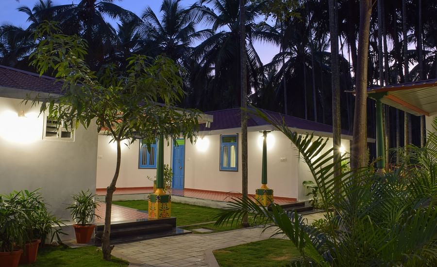 A white resort cottage with a red roof and blue window frames, surrounded by palm trees and green lawns - Ibex Resorts, Coimbatore (Kakarla)