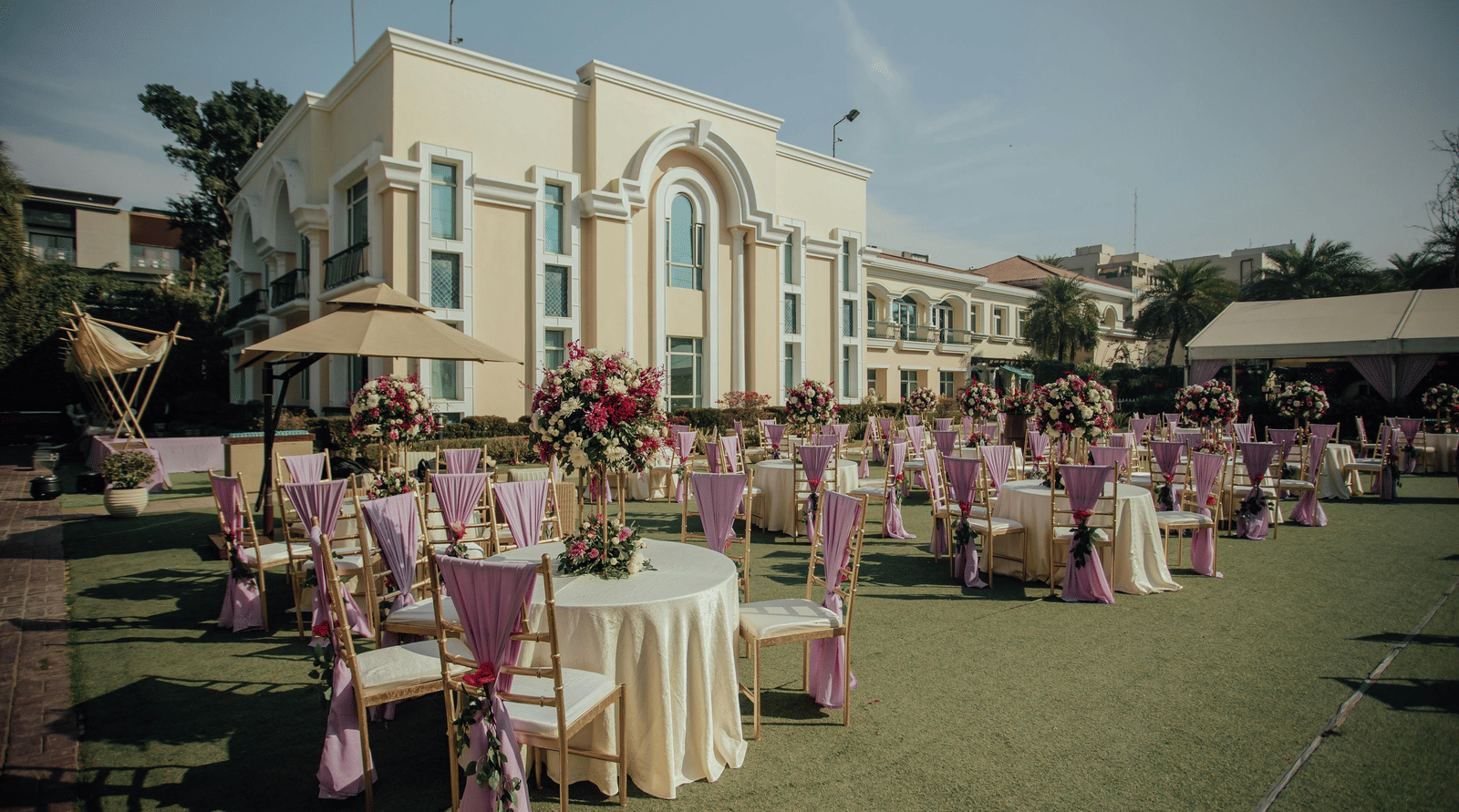 An outdoor wedding setup with round tables, floral decor, and a building in the background.