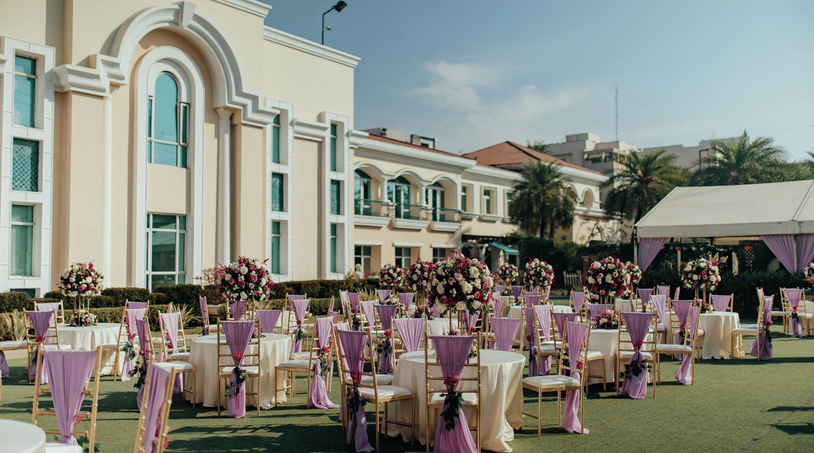 An outdoor event setup with rows of tables and chairs decorated on a lawn in front of a large building.