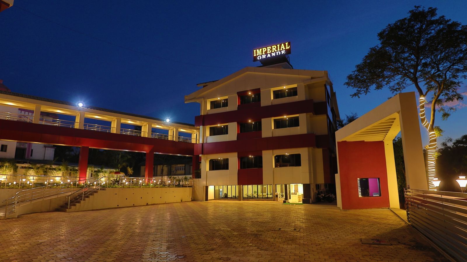 The exterior of Imperial Grande at night, showing a multi-story building with illuminated logo and a paved courtyard reflecting the lights.