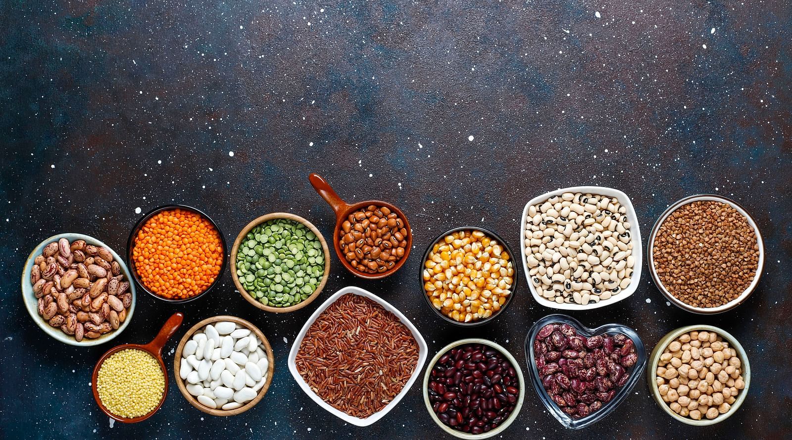 An overhead view of multiple bowls containing assorted spices and seeds on a dark background.