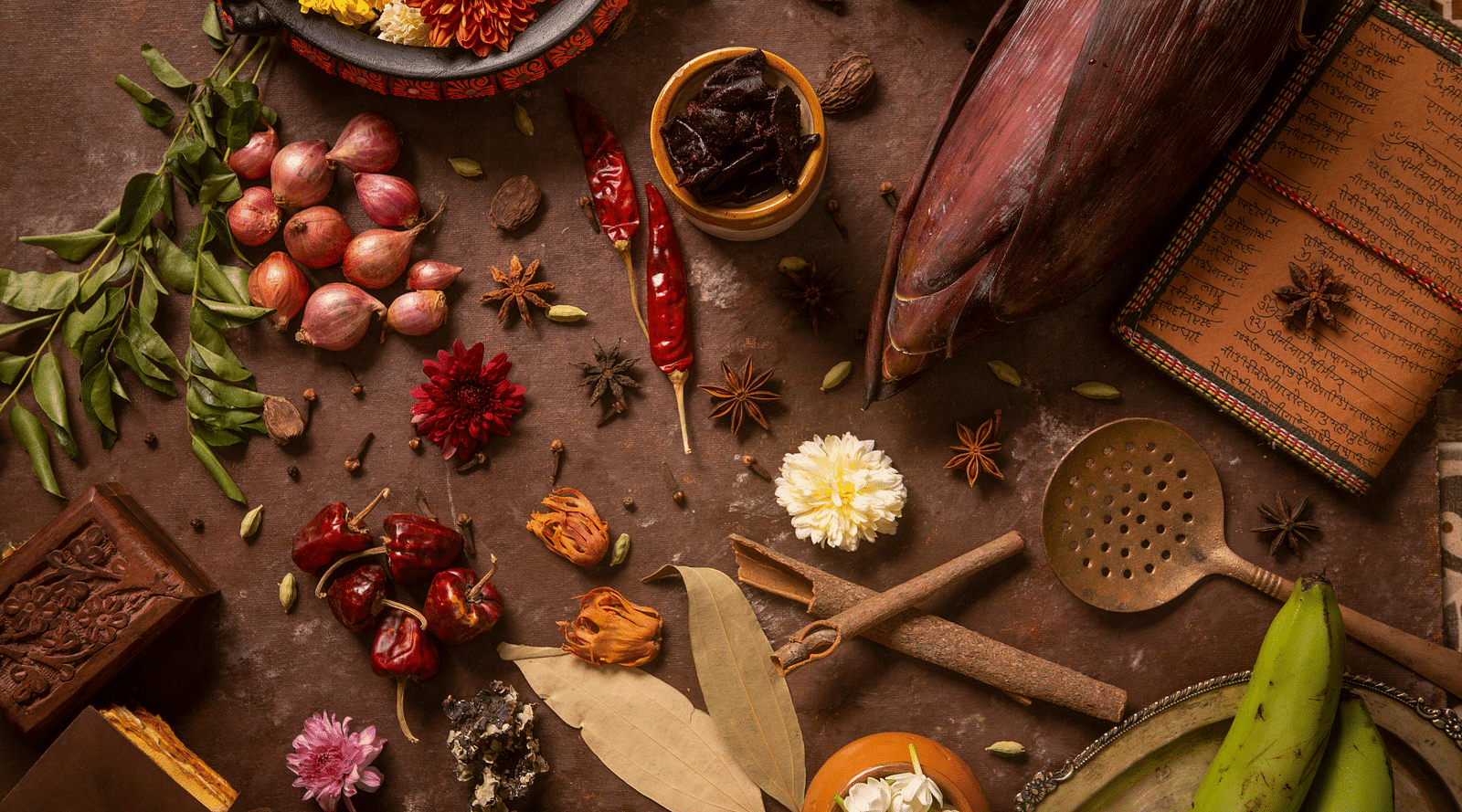 Assorted spices and kitchen tools beautifully arranged with flowers and vegetables on a rustic background | Kamats India