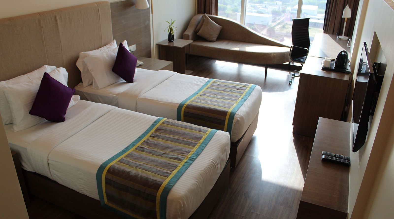 An image showing a pair of twin beds with lush decors, a long sofa with cushion, and a flat screen TV.
