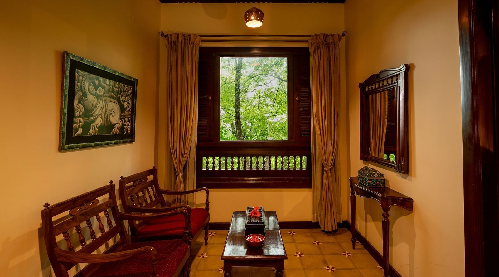 An image of a seating area with wooden chairs, a centre table and an open window  - Hoysala Village Resorts, Hassan