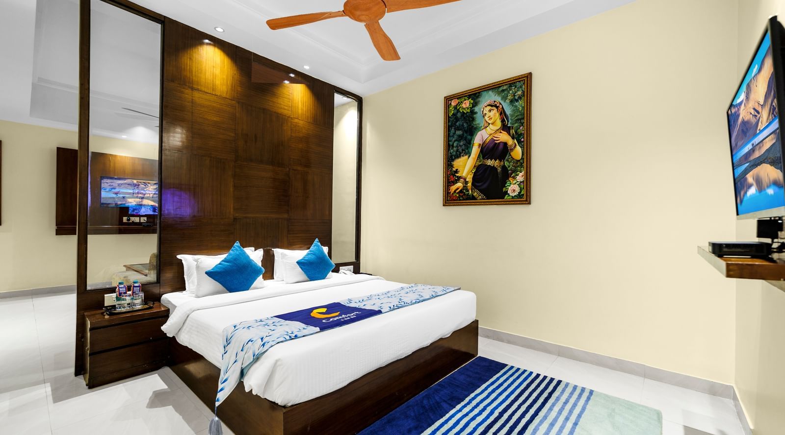 Bedroom at Inde Reva Resorts Rishhikesh with a double bed, blue cushions, ceiling fan, a T.V., and framed artwork on the wall.