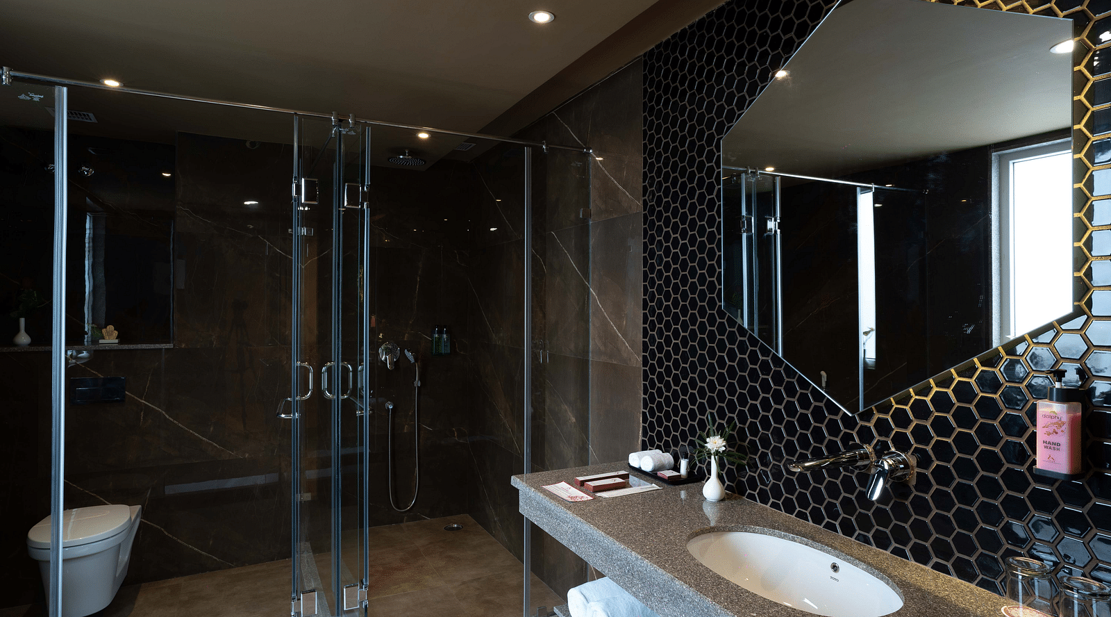 Hexagon-shaped mirror above the washbasin with transparent glass divider separating the showering and toilet areas in the Deluxe room's bathroom at Inde Signature Jaipur.