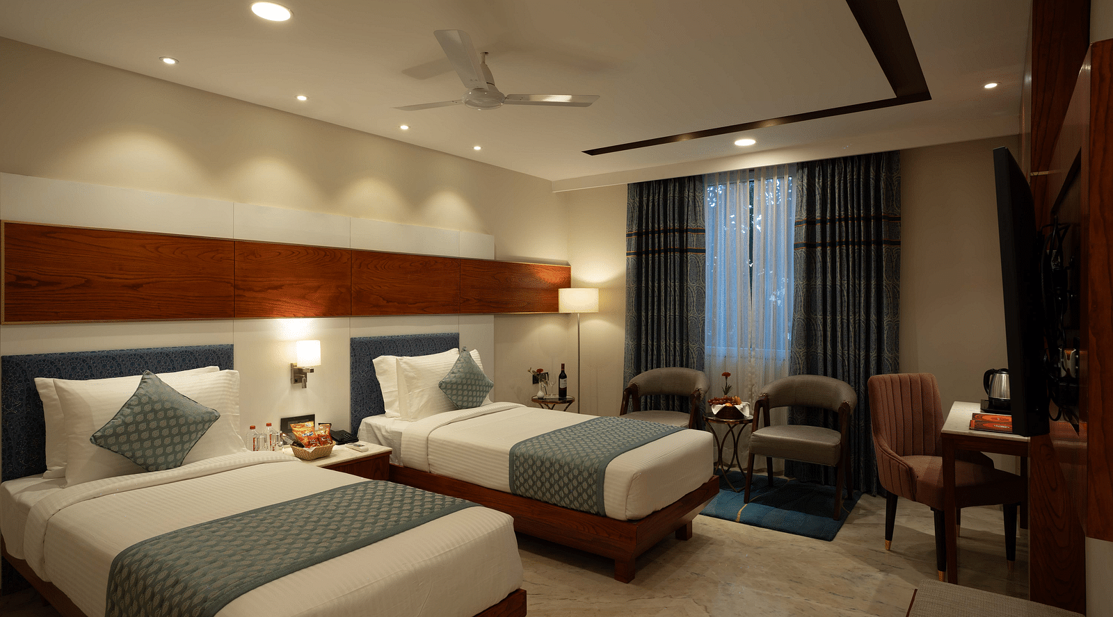 Deluxe Room with twin beds along with a wooden side table with a lamp, wooden table and chair, and two cushioned pieces of furniture at Inde Signature Jaipur