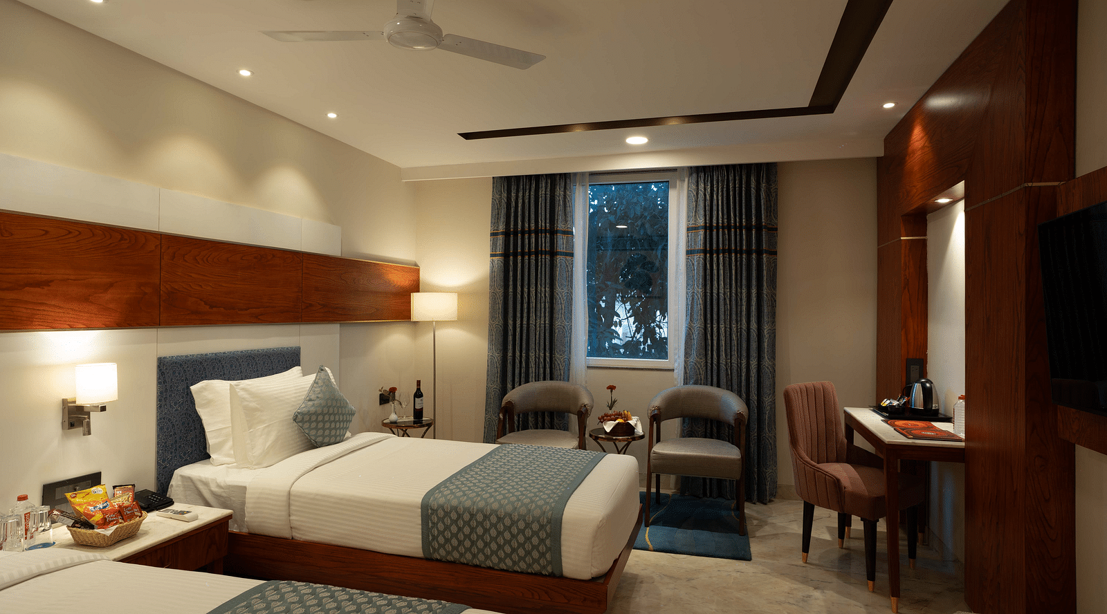 Deluxe Room with twin beds along with a wooden side table with a lamp, wooden table and chair, and two cushioned pieces of furniture at Inde Signature Jaipur