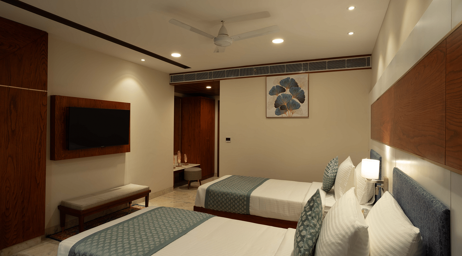 Deluxe Room with twin beds along with a wooden side table with a lamp, wooden table and chair, and wall mounted television at Inde Signature Jaipur