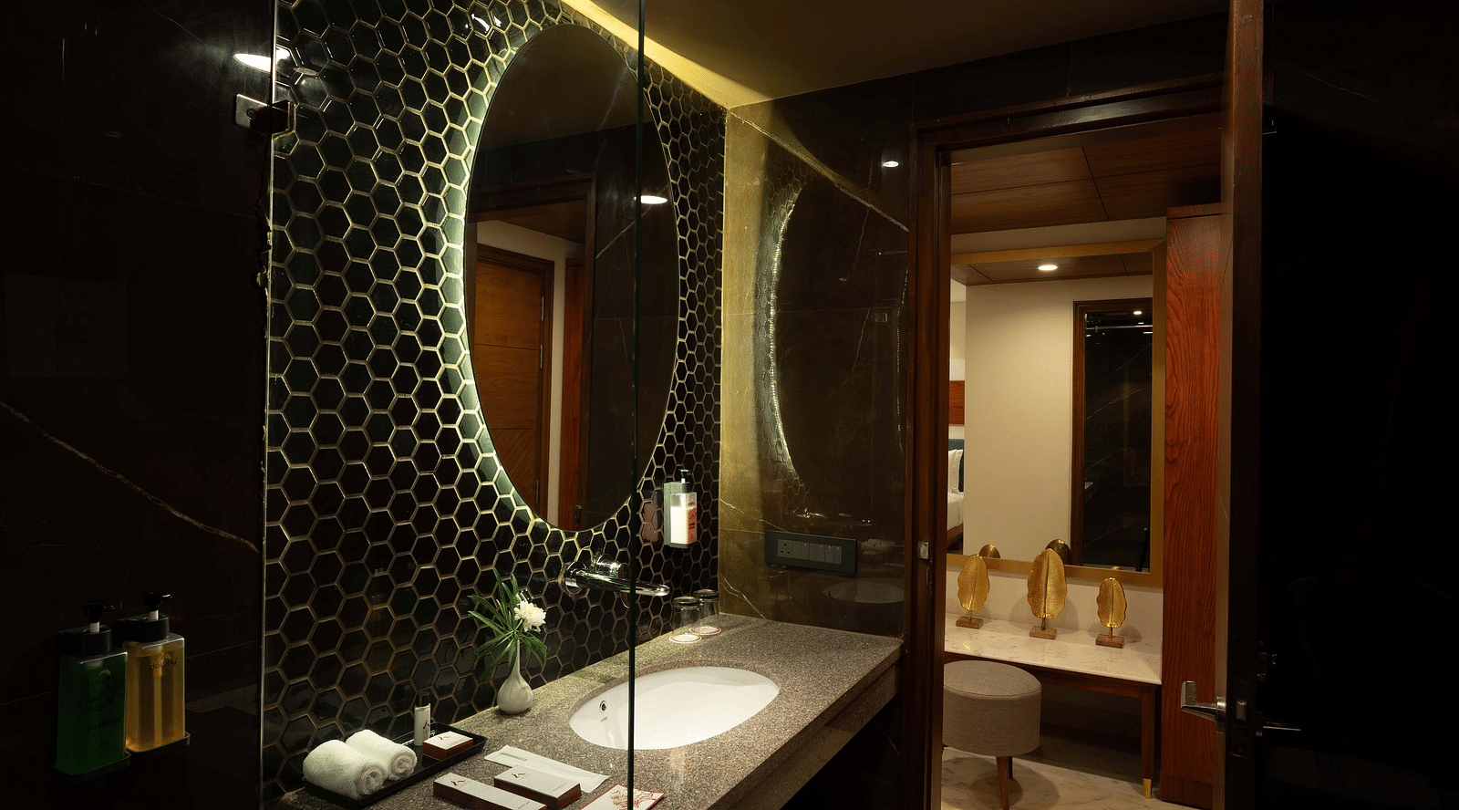 Round shaped mirror above the washbasin with transparent glass divider separating the showering and toilet areas in the Deluxe room's bathroom at Inde Signature Jaipur.