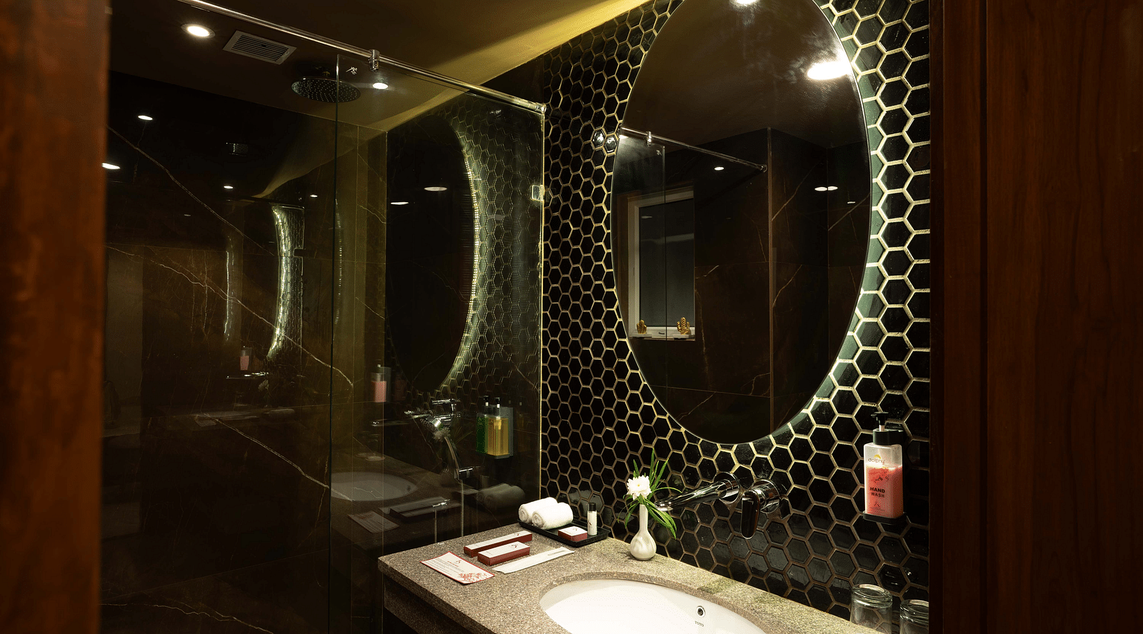Round shaped mirror above the washbasin with transparent glass divider separating the showering and toilet areas in the Deluxe room's bathroom at Inde Signature Jaipur.
