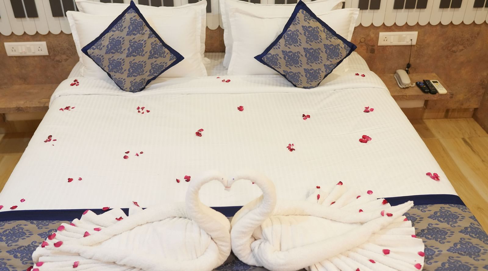 Romantic hotel room setup with white bedding decorated with scattered red rose petals, blue patterned decorative pillows, and swan-shaped towel art at SeaFORT Beach Resort