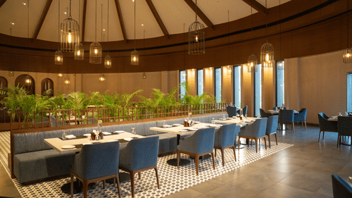 A stylish indoor dining area with blue chairs, round tables, and a high wooden ceiling under warm lighting at Ananta Spa and Resort, Ajabgarh.