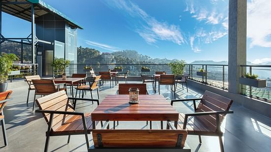 Outdoor dining area at Suryavilas Luxury Resort and Spa, featuring wooden tables and chairs on a spacious terrace with panoramic views of the Himalayan hills under a bright blue sky.
