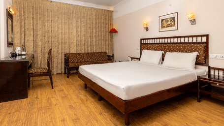 A room with a queen size bed, light wood floors, a work desk, seating area, and floor-to-ceiling beige curtains at Hotel Horizon, Trivandrum.