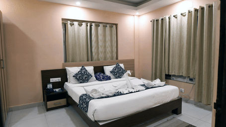 Bedroom of an Executive Suite at Pipul Hotel & Resort, Puri, featuring a plush bed with decorative towel art, patterned cushions, a nightstand, and draped windows.