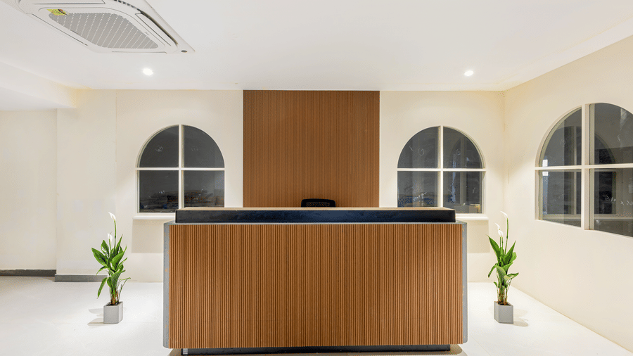 A wooden reception desk, in a white lobby area flanked by potted plants and arched window frames at Monday Hotels & Suites Madhapur Hyderabad.