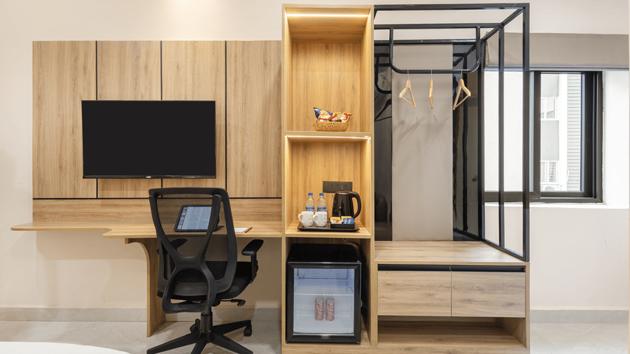 A wooden wall setup featuring a TV screen, a study desk with an office chair, a mini-fridge, and clothing racks at Monday Hotels & Suites Madhapur Hyderabad.