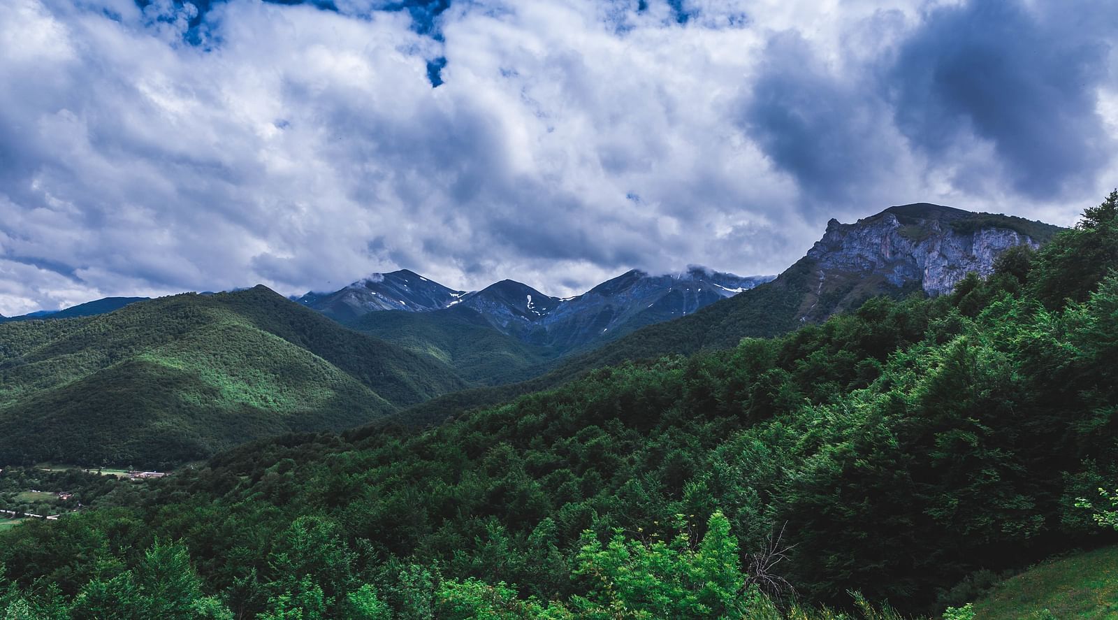 A landscape photo of a mountain range covered in trees, with a dramatic sky filled with clouds.