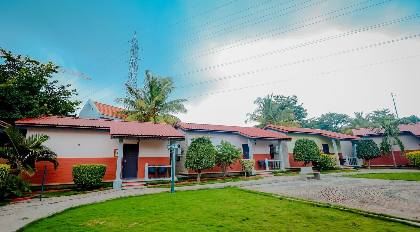 Exterior view of cottages with red roofs and green lawns at Nirantara Resort & Spa, Bengaluru, under a partly cloudy sky.