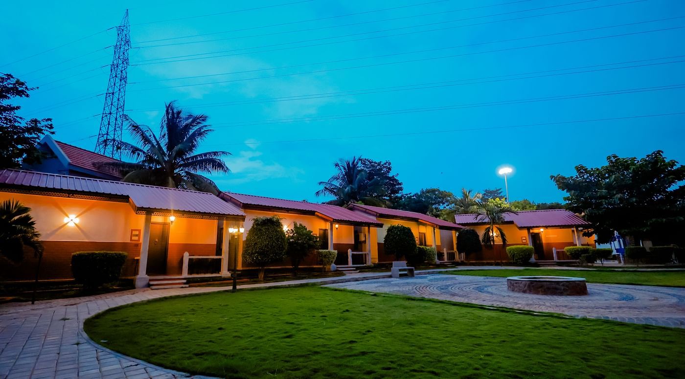 Exterior view of cottages with red roofs and green lawns at Nirantara Resort & Spa, Bengaluru, at dusk with lights illuminating the pathways.