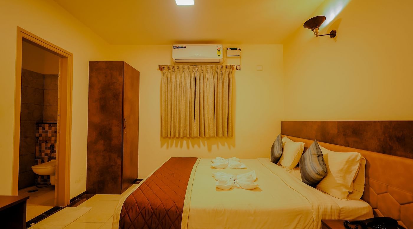 Side angle view of a room at Niratara Resort and Spa featuring a bed, window with short curtains, an almirah, and the bathroom door.
