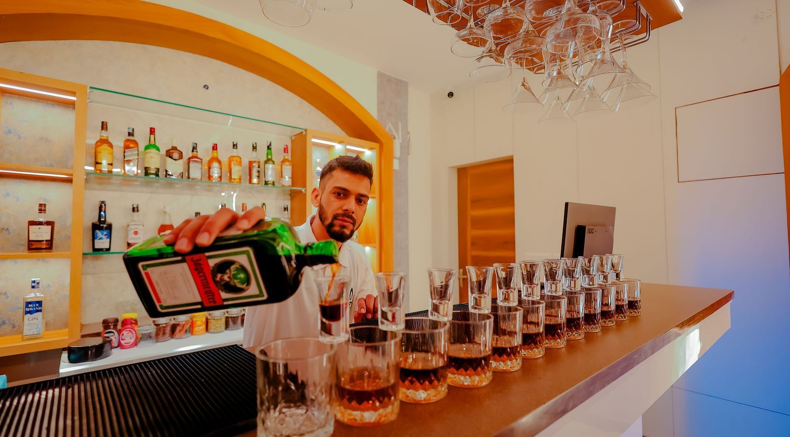 A bartender prepares drinks at a well-stocked bar at Nirantara Resort & Spa, showcasing a variety of liquors and glassware.