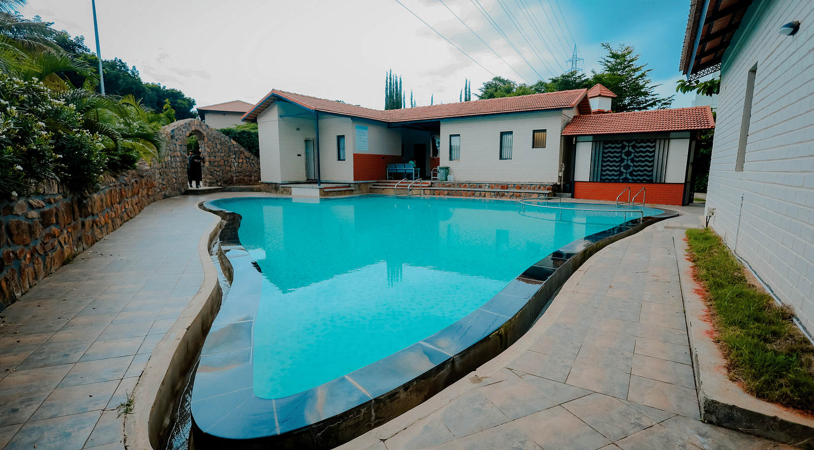 A uniquely shaped swimming pool at Nirantara Resort & Spa, Bengaluru, surrounded by plants and buildings.