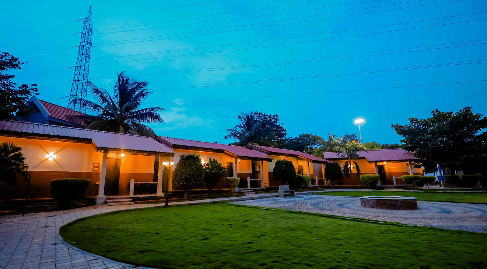 Exterior view of cottages with red roofs and green lawns at Nirantara Resort & Spa, Bengaluru, at dusk with lights illuminating the pathways.