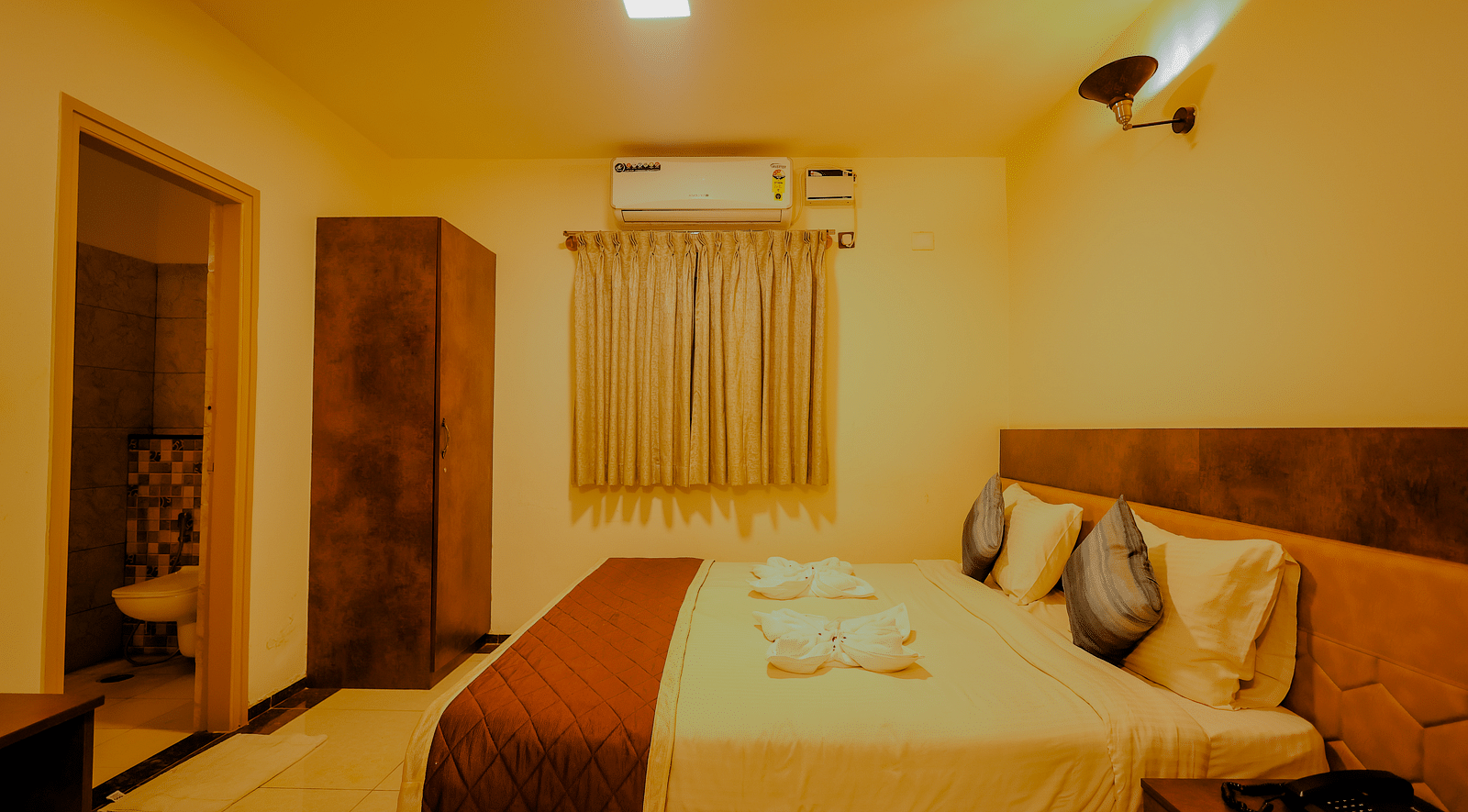 Side angle view of a room at Niratara Resort and Spa featuring a bed, window with short curtains, an almirah, and the bathroom door.