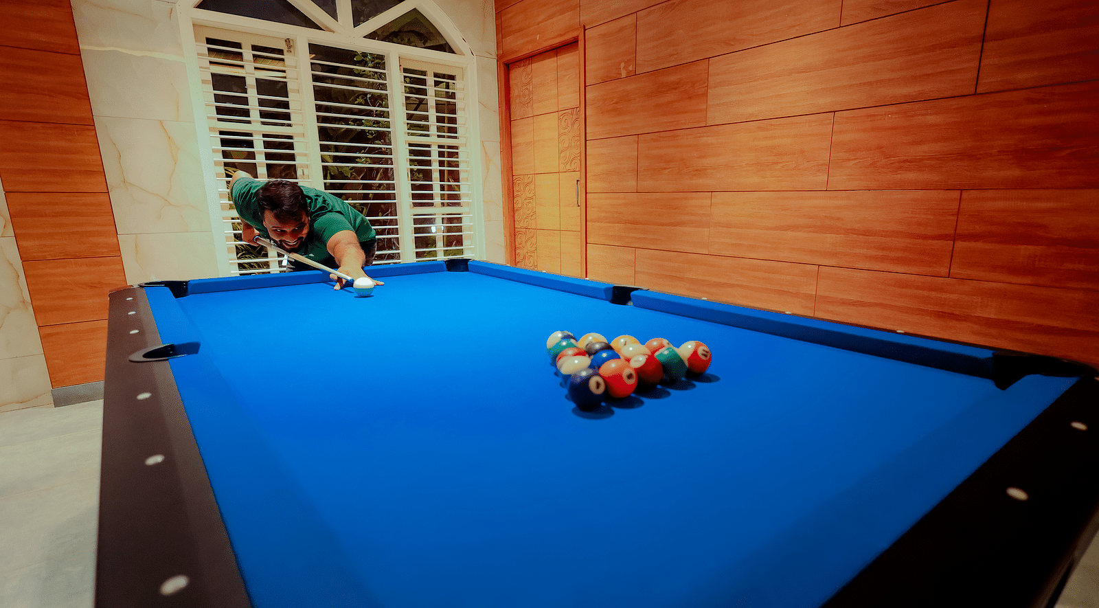 A person playing pool on a blue pool table in a room with a brick wall and arched window at Nirantara Resort & Spa.