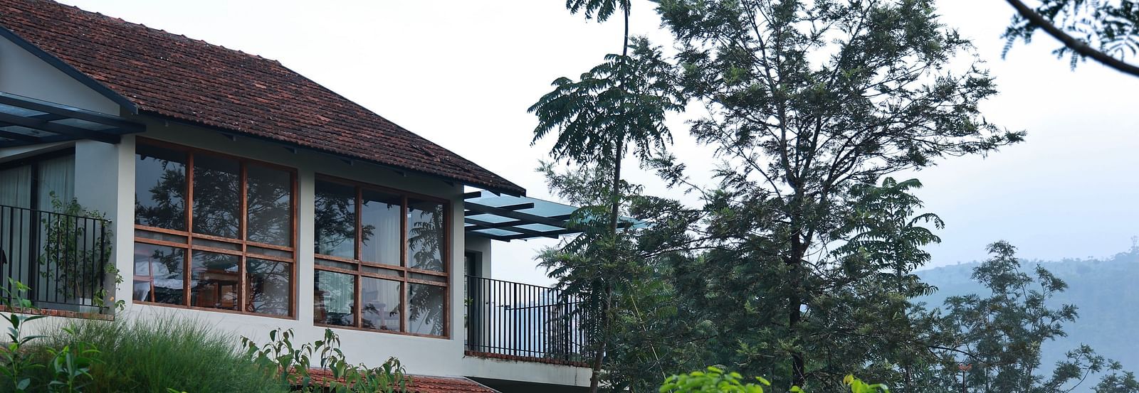  A building at Vaastha Vagamon, nestled amidst abundant foliage, with large windows overlooking a verdant landscape.