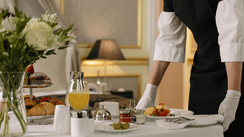 Hotel staff serving in-room breakfast with croissants, juice, flowers, and a plated hot dish on a white table cloth.