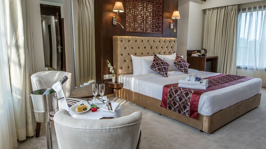 Bedroom at Clarks Inn Suites, Near Gwalior DD Mall, with a king size bed, patterned pillows and runner, wall lamps, two chairs with a table setup, wine cooler, and large windows with curtains.