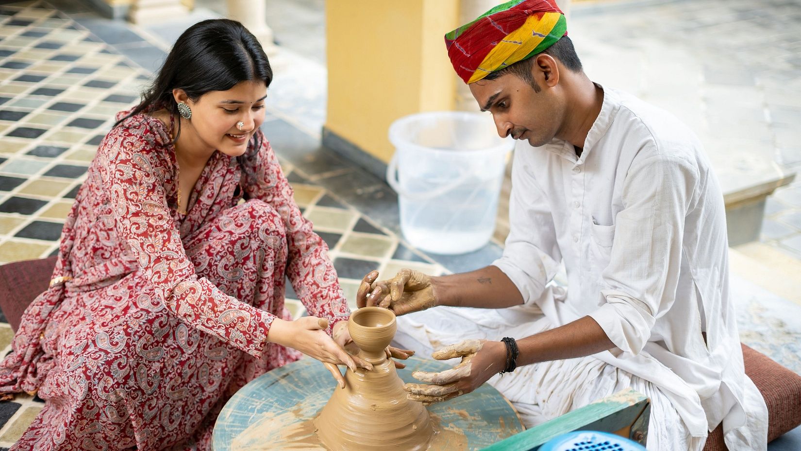 A man teaching pottery to a woman, both focused on shaping clay on a pottery wheel.