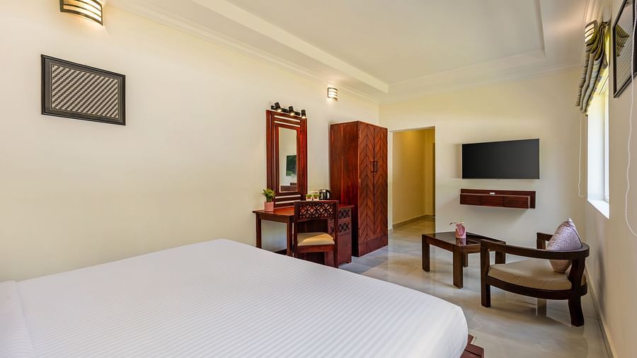 A well-lit hotel room with a bed, a desk, a wardrobe, a television, and a seating area - Mango Hill Resort, Coimbatore