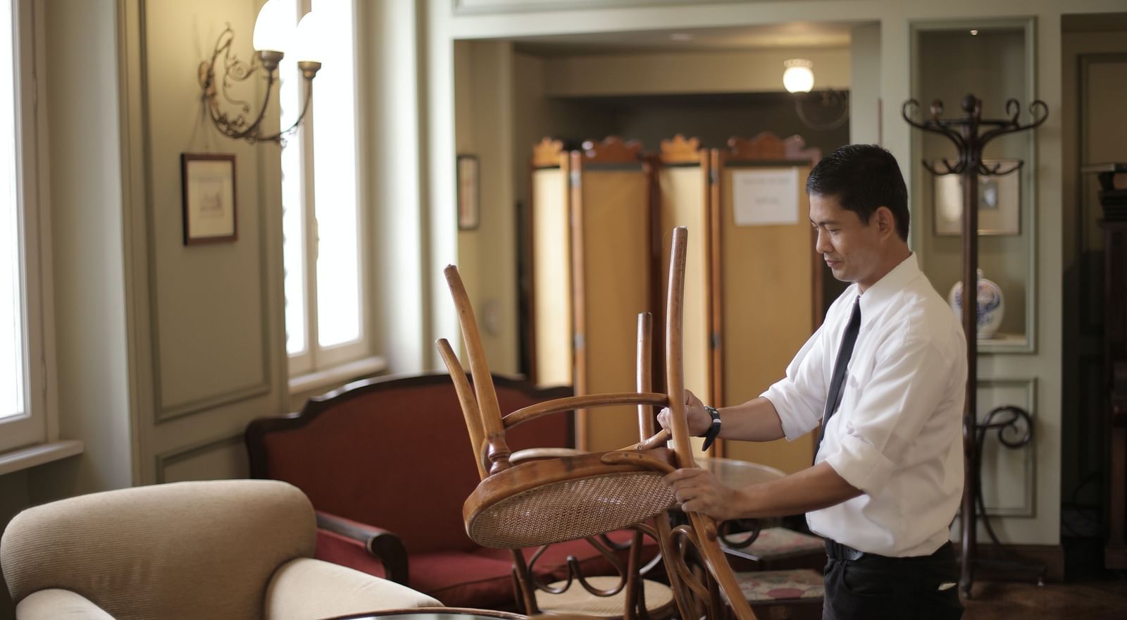 A man in a white shirt and tie setting up chairs in a well-lit room with elegant decor