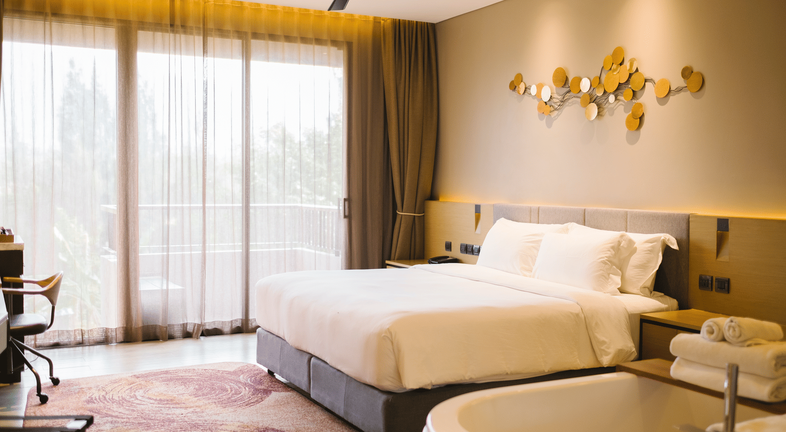 A bright and cosy room featuring a double bed, stylish decor, large windows with balcony access, ceiling fan, and modern furnishings.