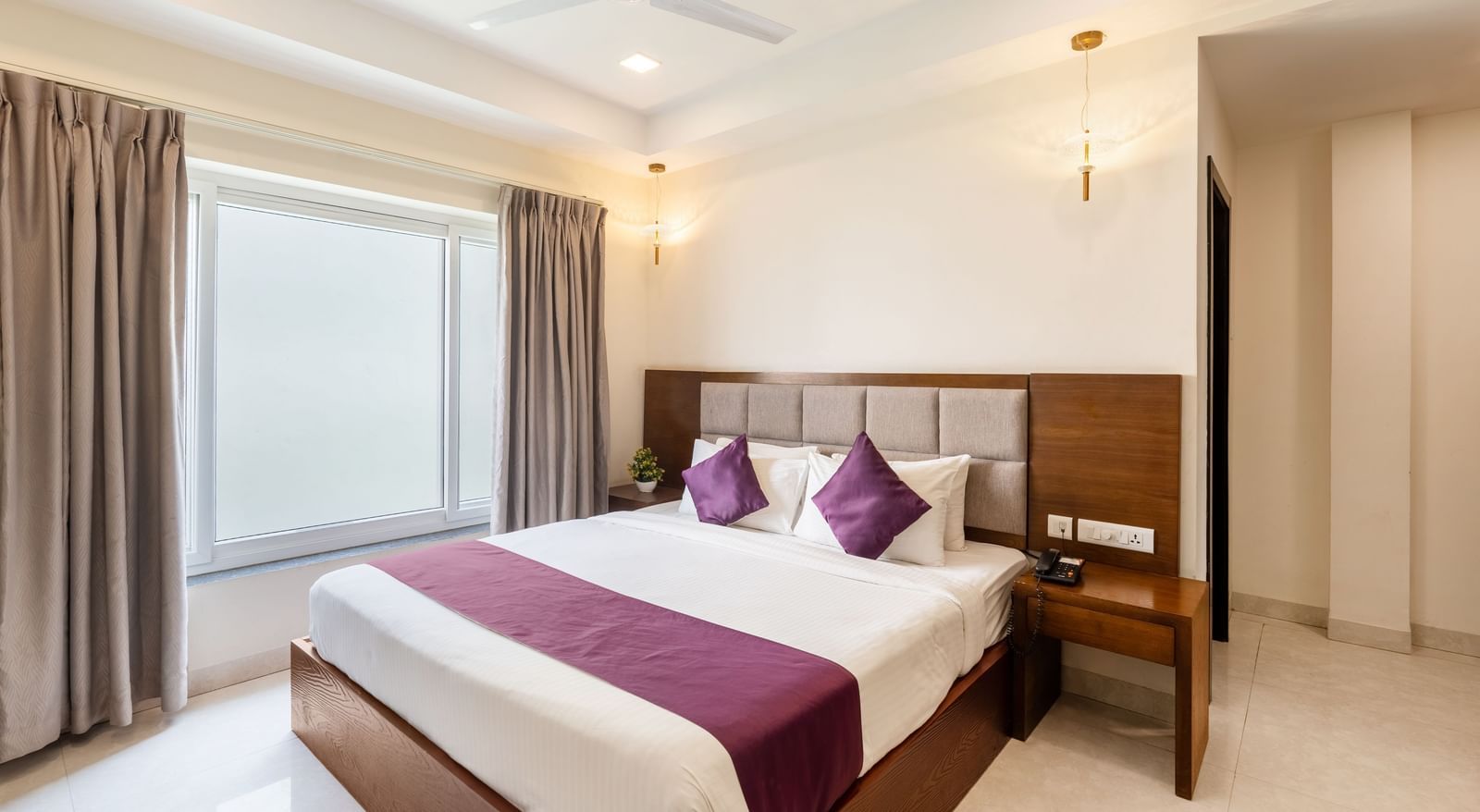 A Premium Room at Destino by Monday Hotels, Gachibowli, with a large double bed with purple accents and a ceiling fan, illuminated by natural light from a large window.